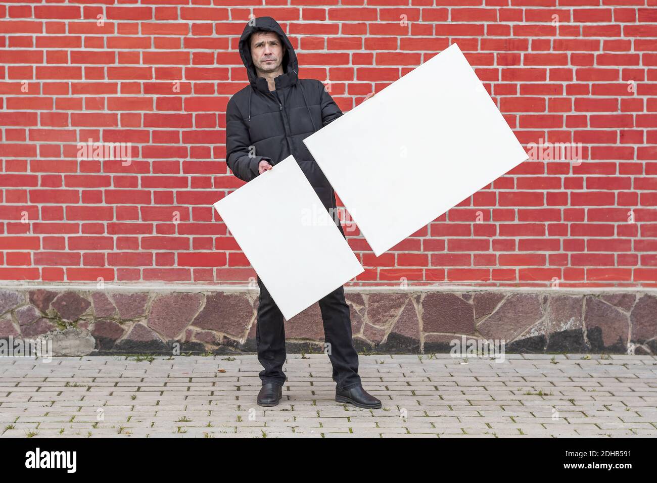 A young man stands against a background of a red brick wall and holds in front of him two white blank canvases for painting Stock Photo