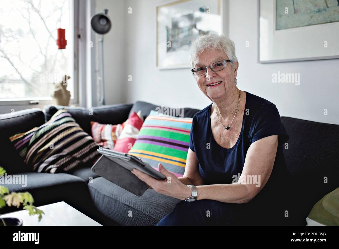 Portrait of confident senior woman sitting with digital tablet on sofa in living room at home Stock Photo