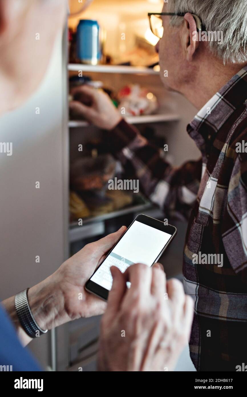 Senior woman holding smart phone standing by man in kitchen at home Stock Photo