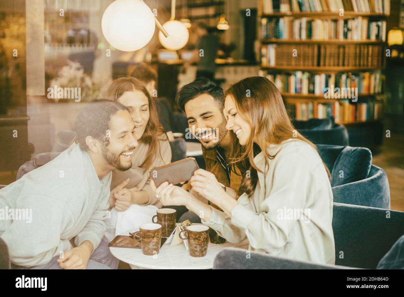 Happy woman showing smart phone to friends while sitting in restaurant seen through glass Stock Photo