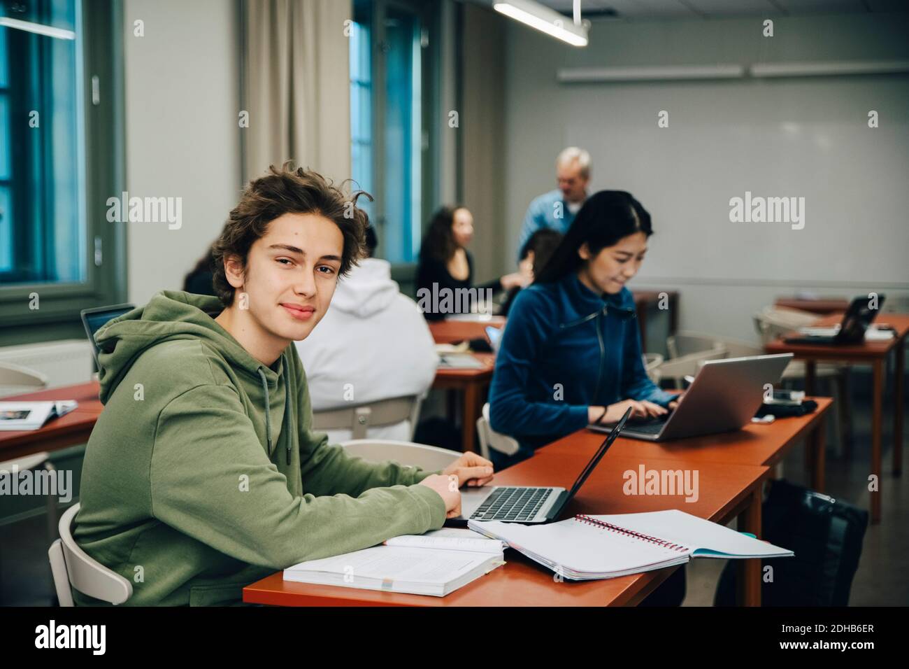 Portrait of smiling teenage boy using laptop at desk with classmates ...