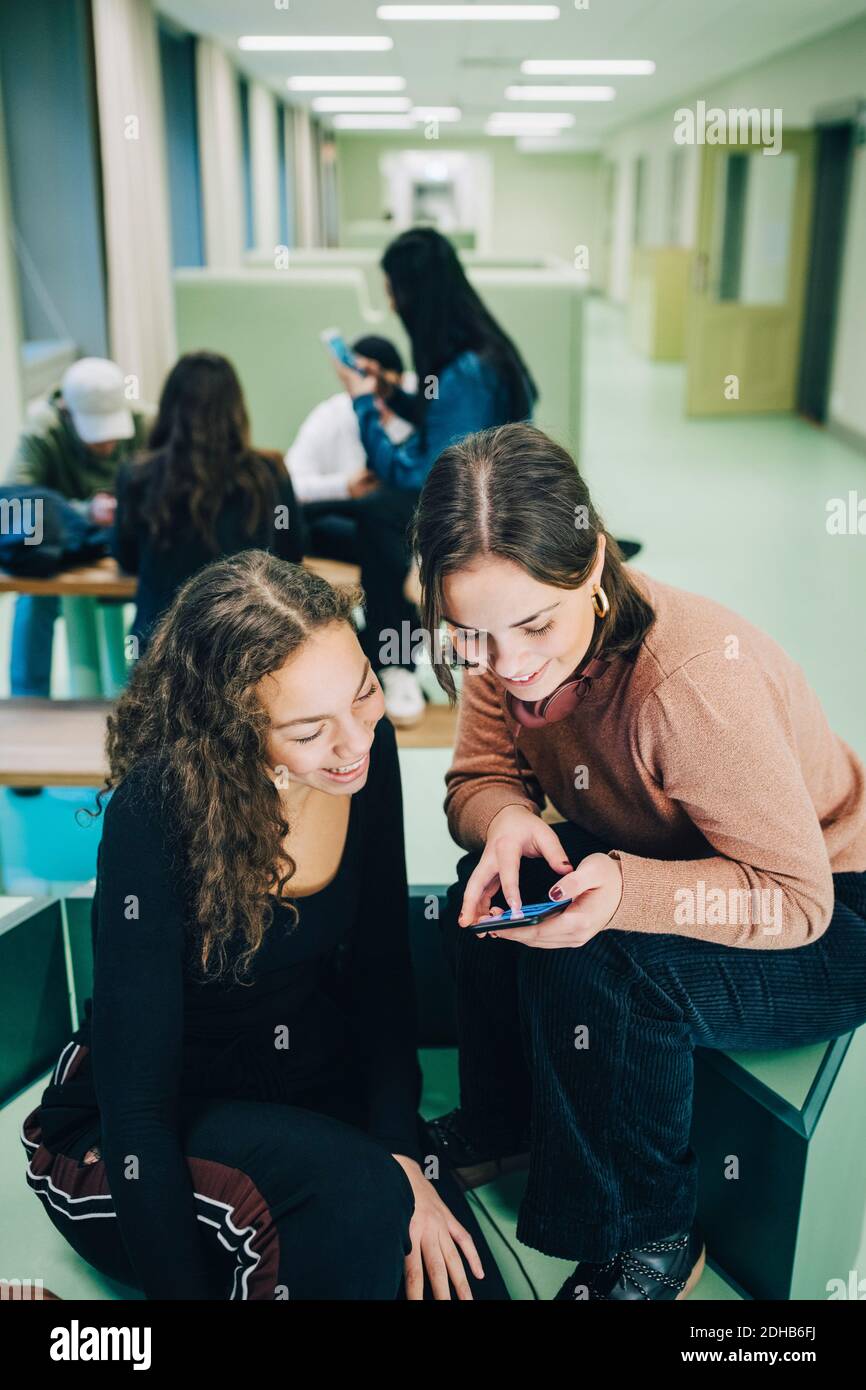 High angle view of teenage girl showing smart phone to female classmate while sitting at corridor in school Stock Photo