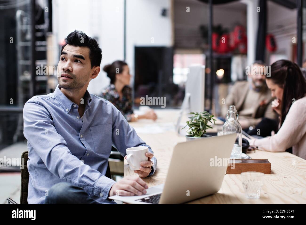 Thoughtful businessman having coffee and using laptop while sitting with colleagues at table Stock Photo