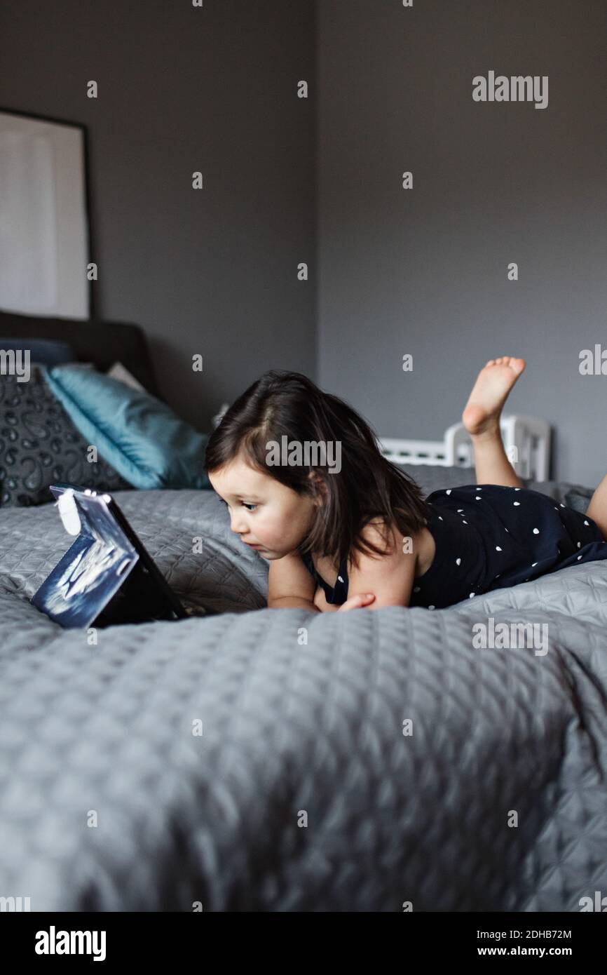 Full length of cute girl watching digital tablet while lying on bed at ...
