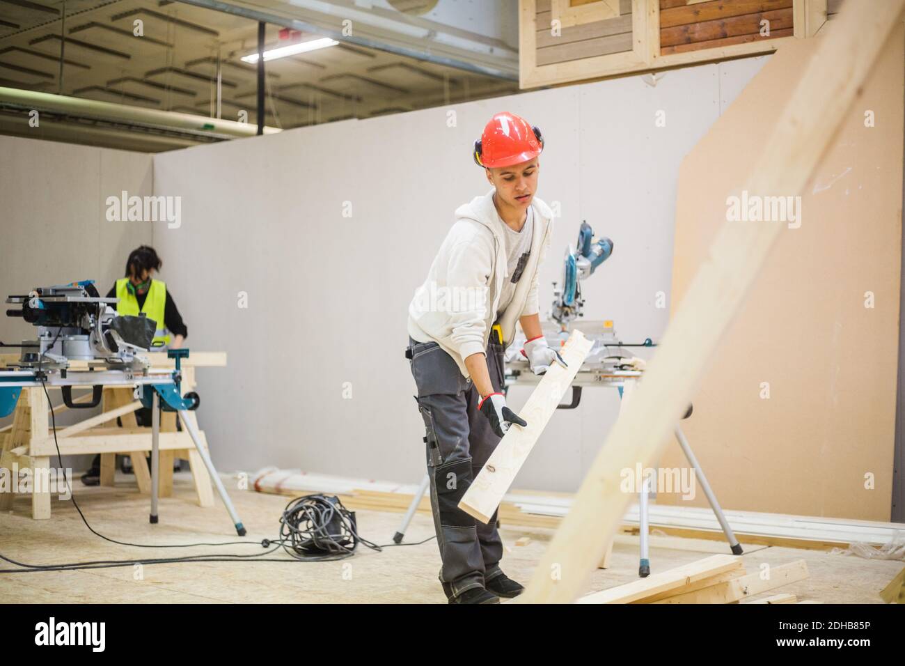 Male teenage carpenter holding plank by electric saw at illuminated workshop Stock Photo