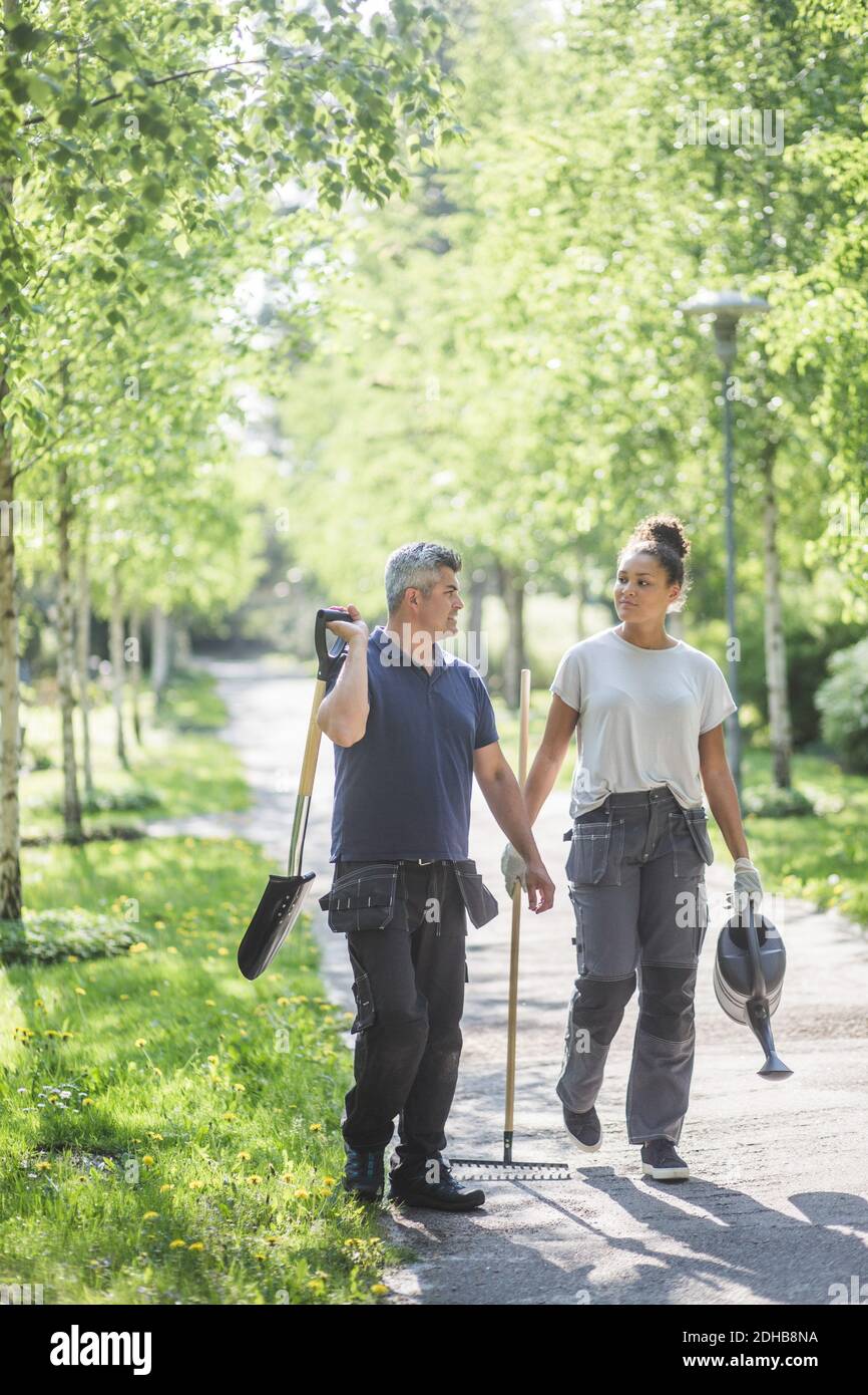 Full length of female trainee with male instructor holding gardening equipment and walking footpath at garden Stock Photo