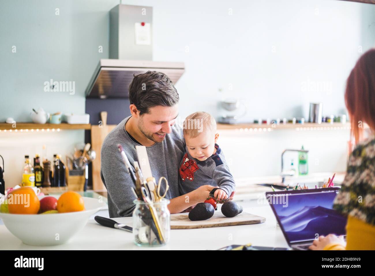 Smiling man carrying son holding tomato on cutting board at kitchen island Stock Photo