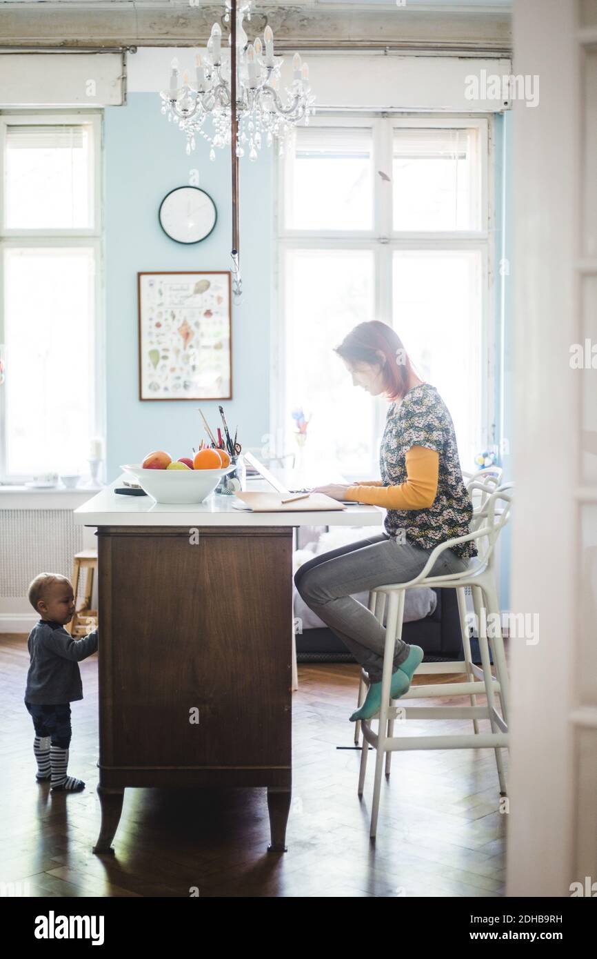 Full length of woman working on laptop with son standing by kitchen island at home Stock Photo