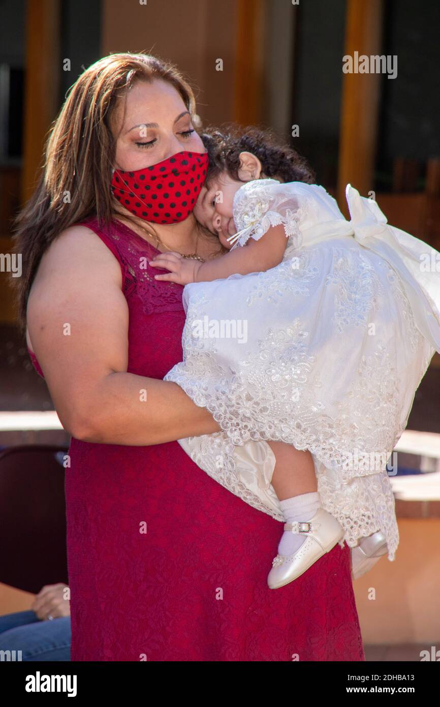 Wearing a face mask due to the coronavirus pandemic, a Hispanic mother brings her daughter at an outdoor christening ceremony at a Southern California Stock Photo