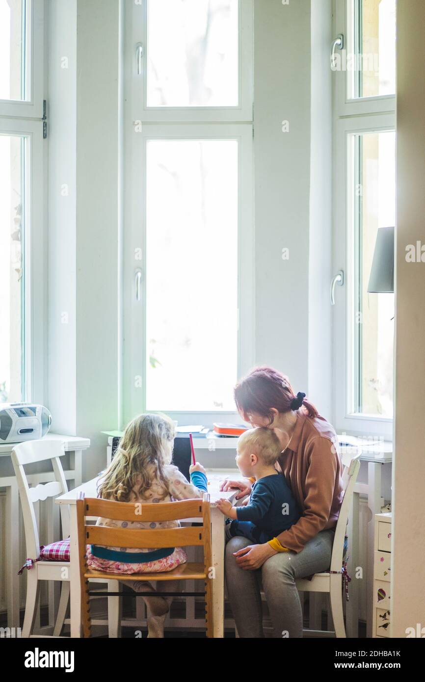 Mother sitting with son while assisting girl studying at table Stock Photo