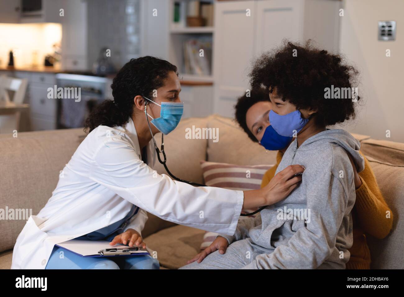 Mixed race girl wearing face mask being examined by mixed race female doctor sitting on couch. self isolation at home together during coronavirus covi Stock Photo