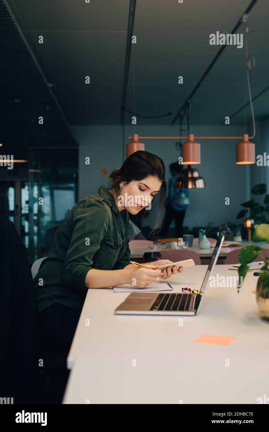 Side view of businesswoman using smart phone while working at desk in creative office Stock Photo