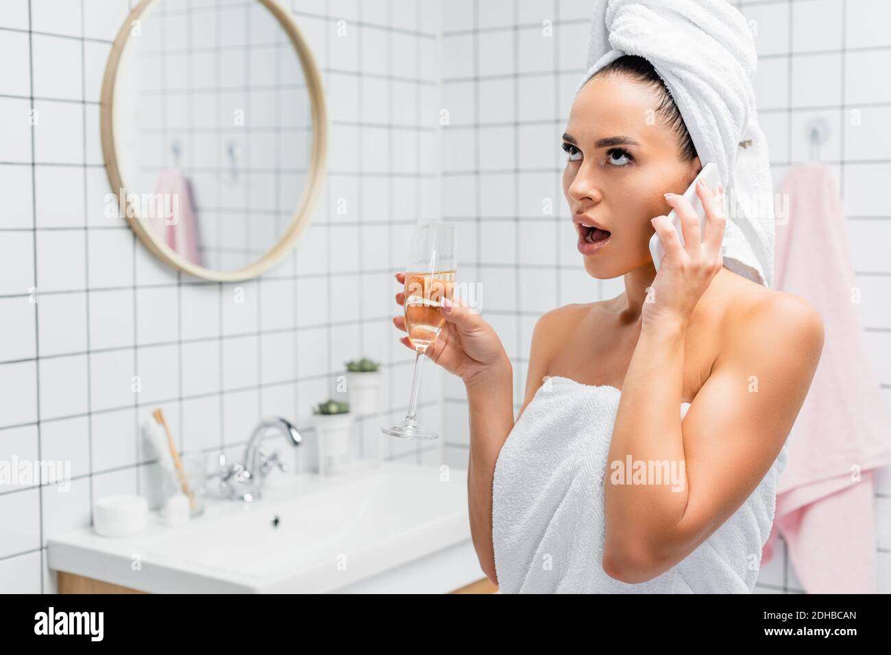 Shocked woman in towels talking on smartphone while holding glass of