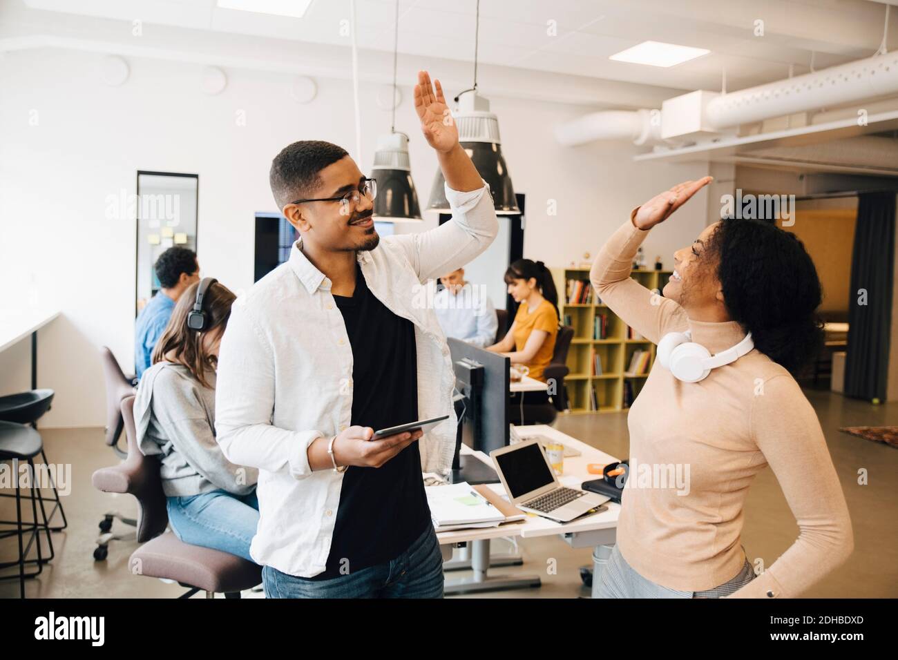 Smiling programmers doing high-five while standing with colleagues working in background Stock Photo
