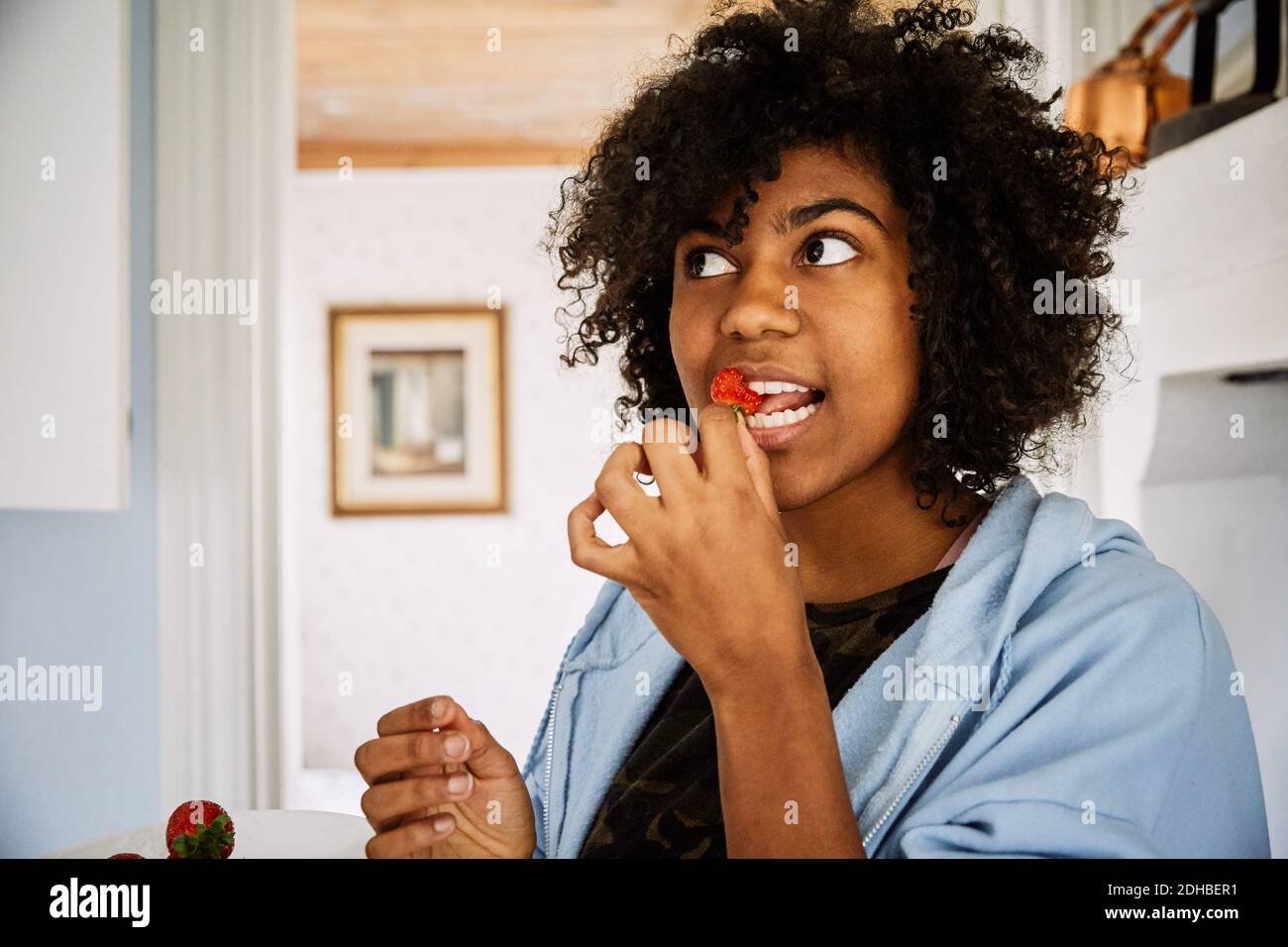 Young woman with curly hair eating strawberry while standing at home Stock Photo