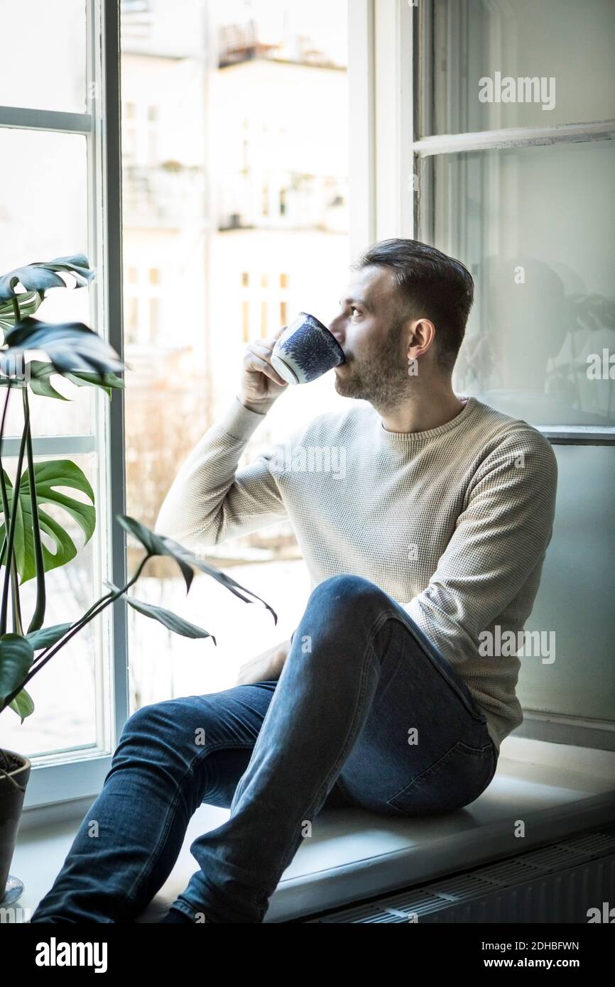 Thoughtful young man drinking coffee while sitting on window sill Stock Photo