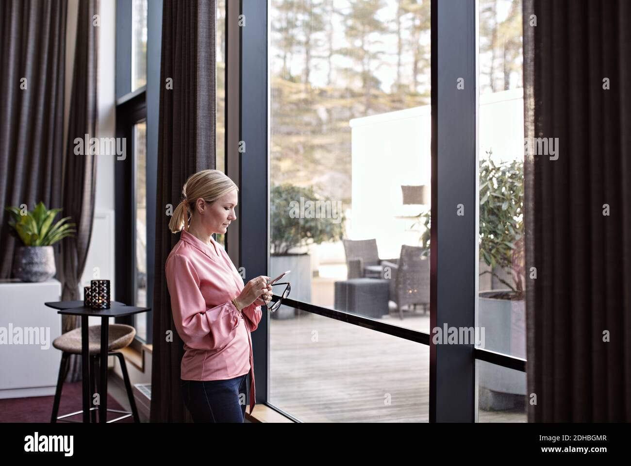 Businesswoman using phone while standing by window in office Stock Photo