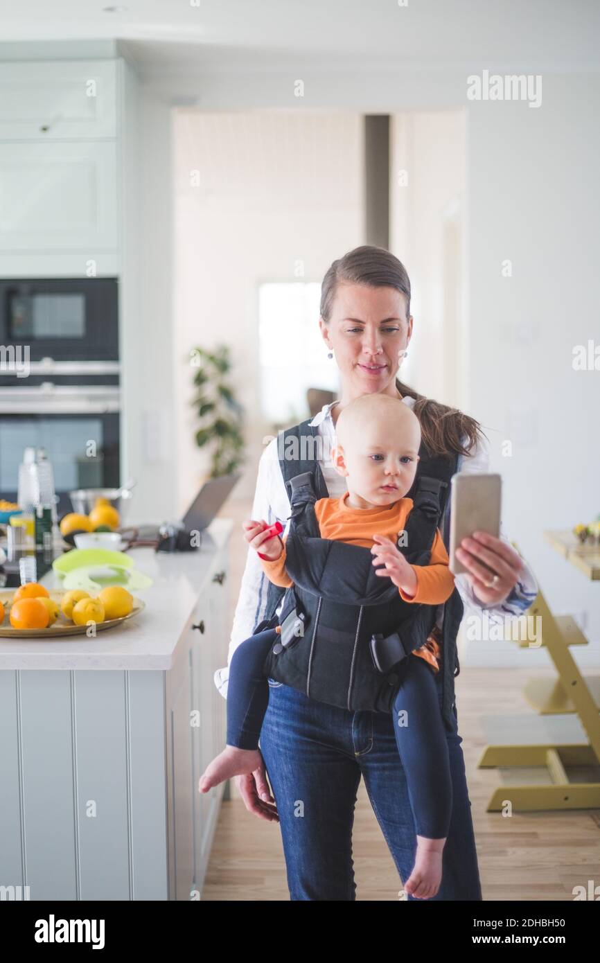 Female freelancer blogging while carrying daughter in kitchen using smart phone at home Stock Photo