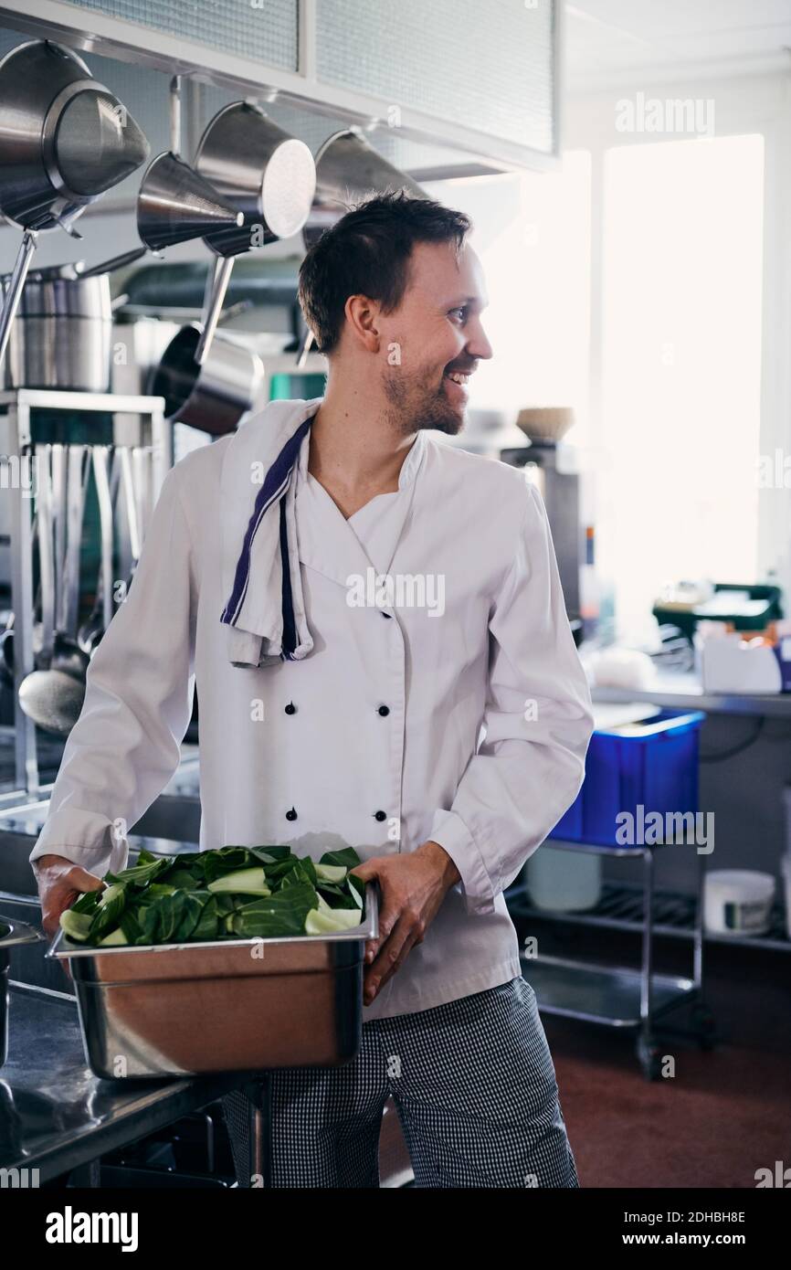 Young male chef smiling while holding container of leaf vegetable in kitchen Stock Photo