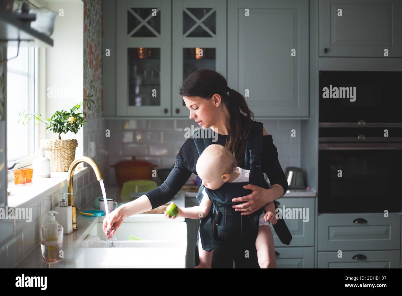 Mother washing hand in kitchen sink while carrying baby girl at home Stock Photo