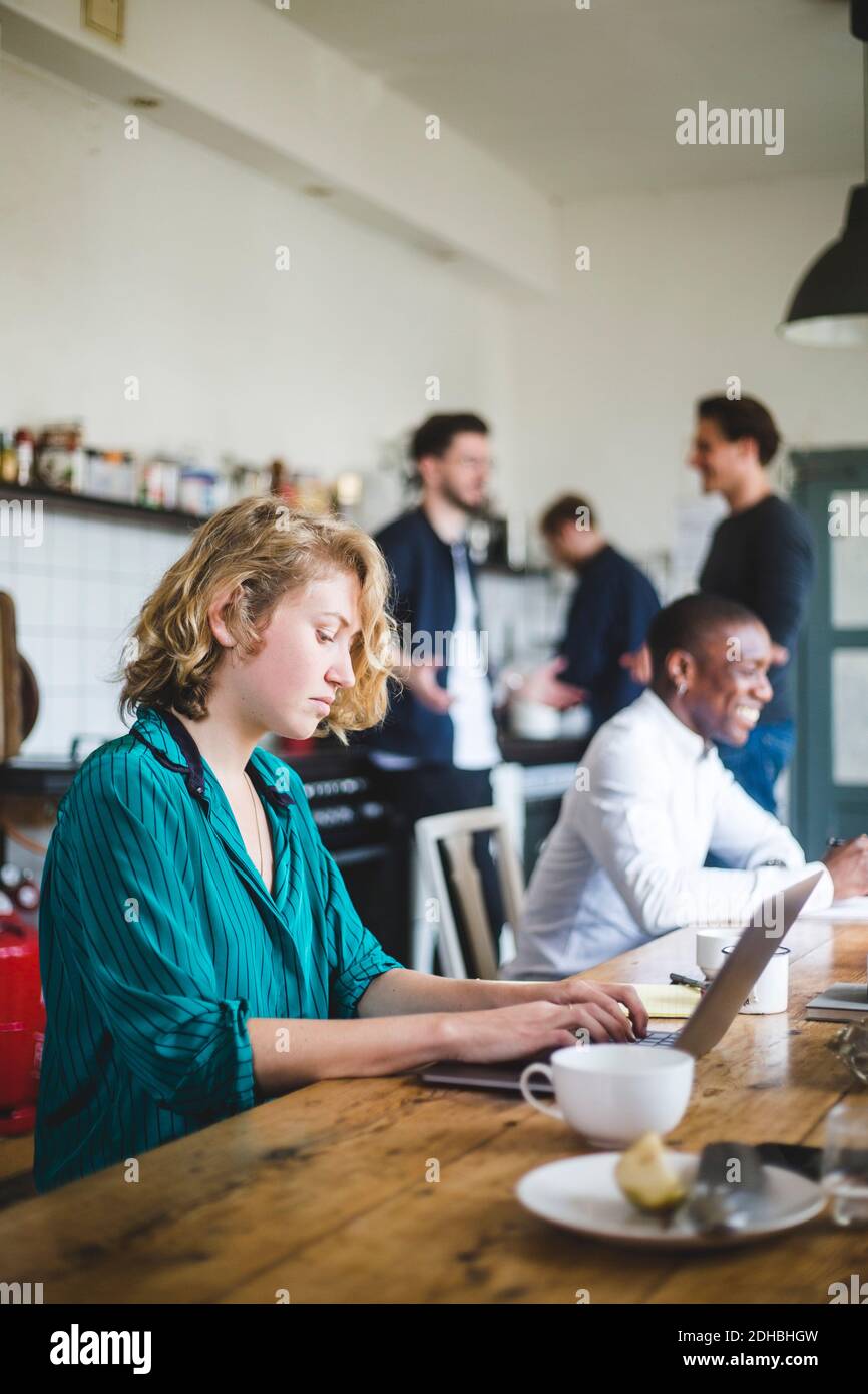 Serious female IT hacker using laptop while colleagues talking in background at creative workplace Stock Photo