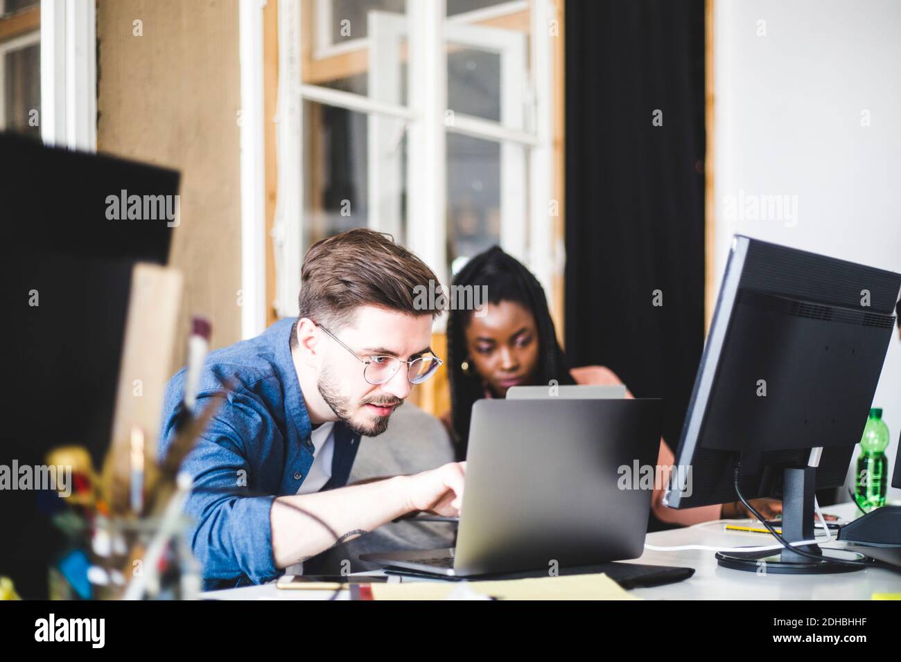 Young computer expert coding in laptop while working with female colleague at workplace Stock Photo
