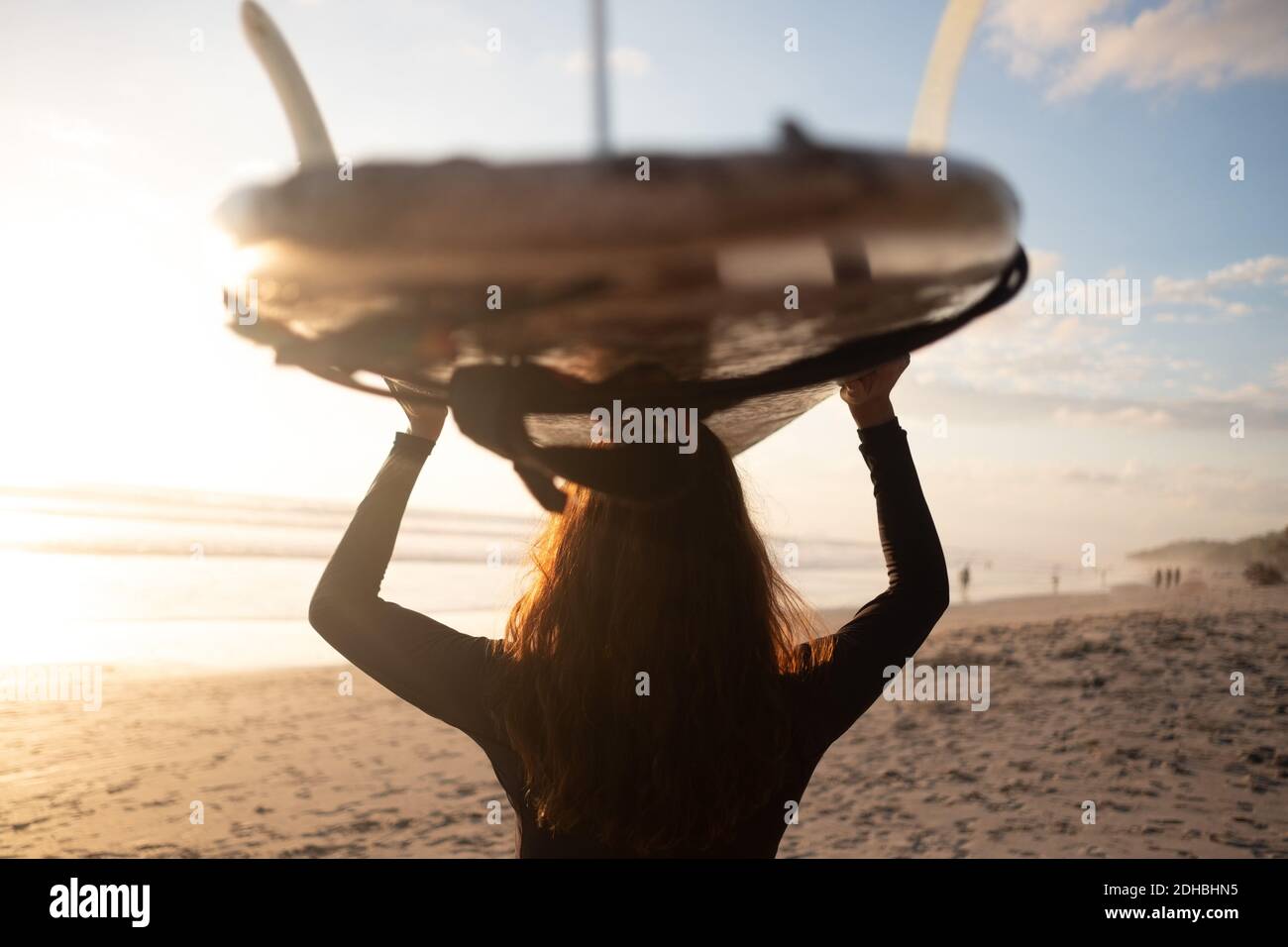 Rear view of mature woman carrying surfboard on head at beach Stock Photo