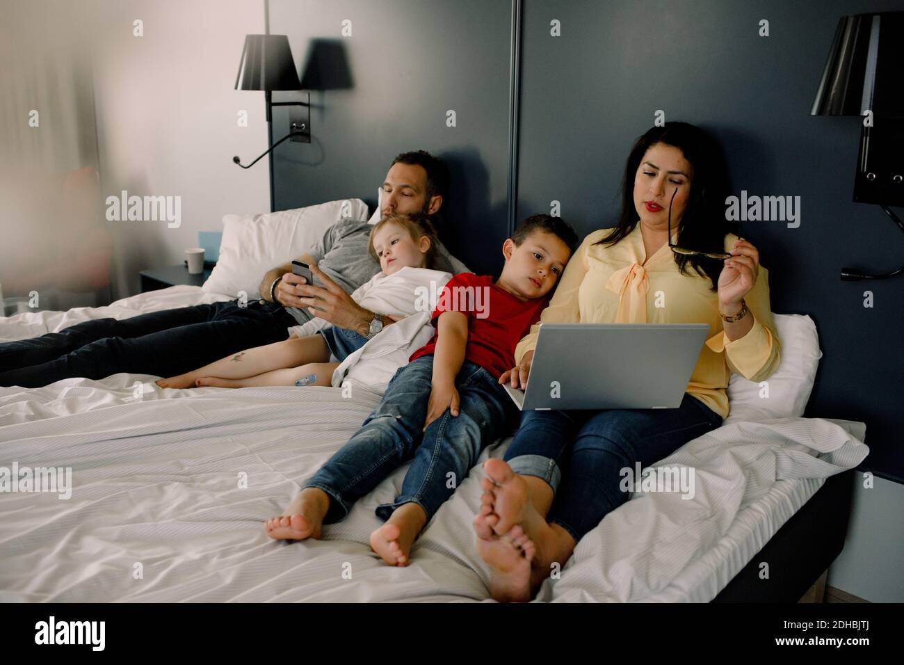 Parents using technologies while sitting with children on bed in bedroom Stock Photo