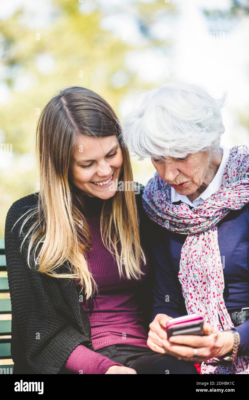 Senior woman showing mobile phone to granddaughter while sitting on bench outdoors Stock Photo