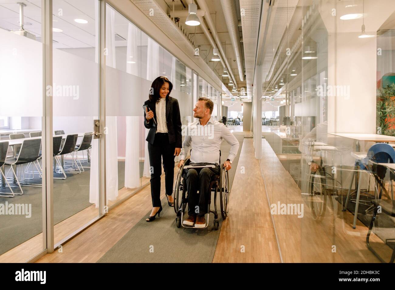 Smiling businesswoman looking at disabled colleague sitting on wheelchair in corridor at work place Stock Photo