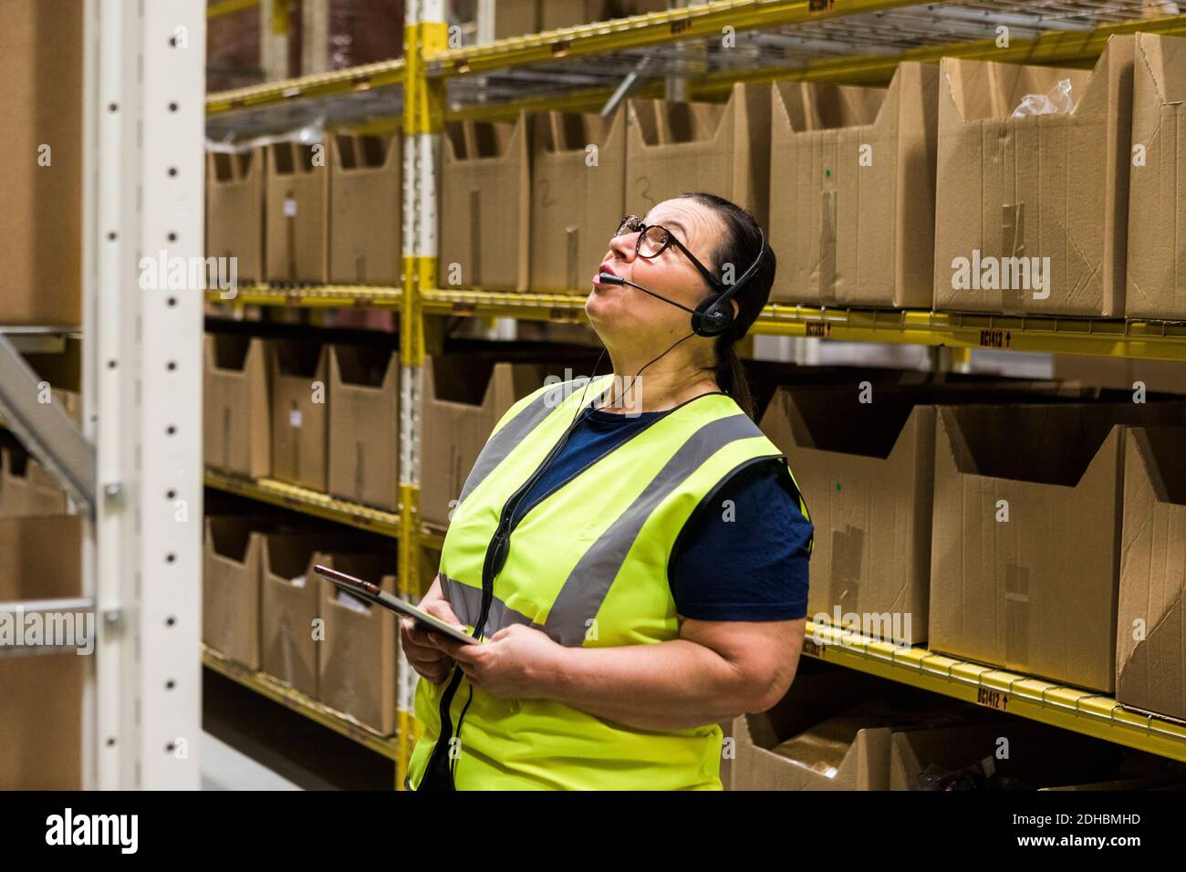 Mature female worker with digital tablet looking up while talking on headset against rack at distribution warehouse Stock Photo