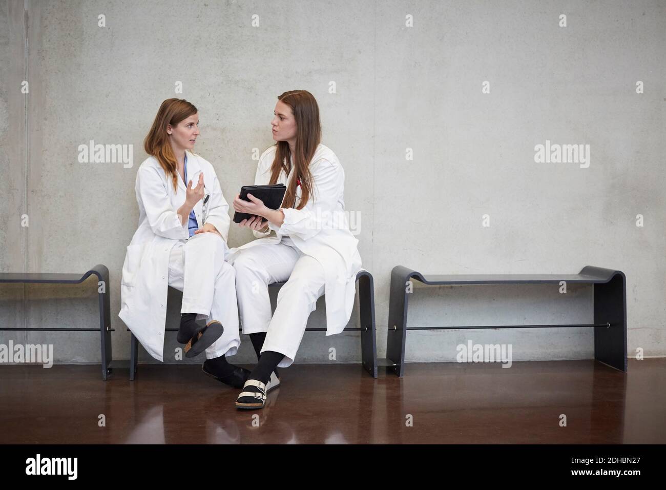 Full length of confident female doctors discussing over digital tablet while sitting against wall at hospital Stock Photo