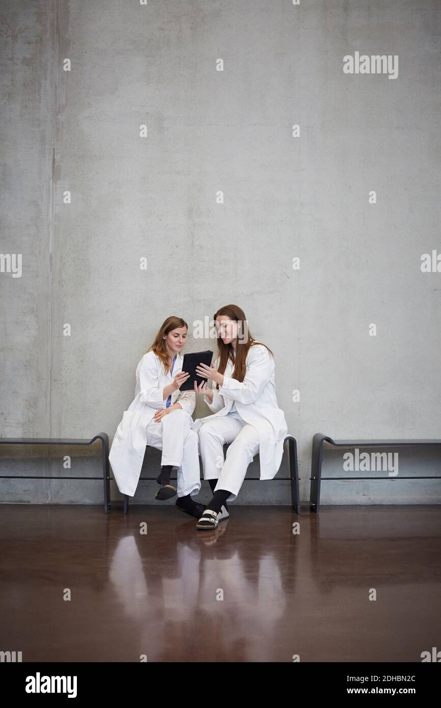 Full length of female doctors discussing over digital tablet while sitting against wall at hospital Stock Photo