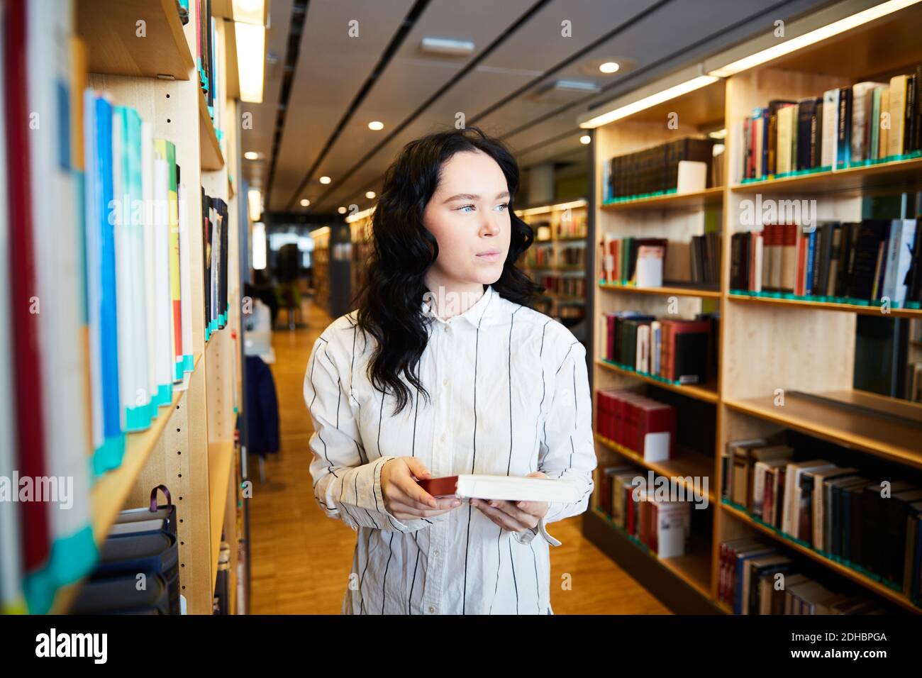 Young female student holding book while standing in university library Stock Photo