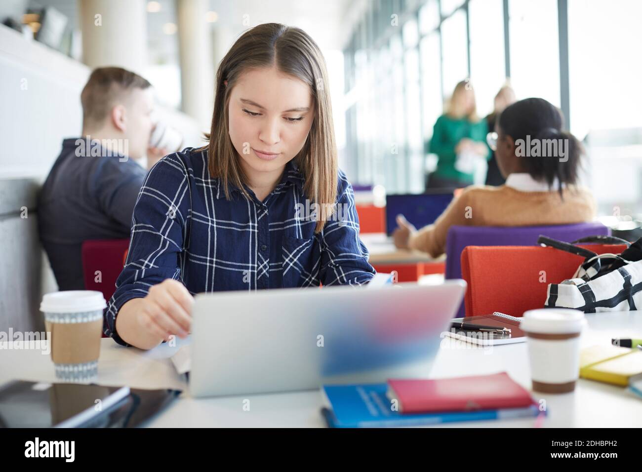 Female student using laptop at table with friends in background Stock Photo