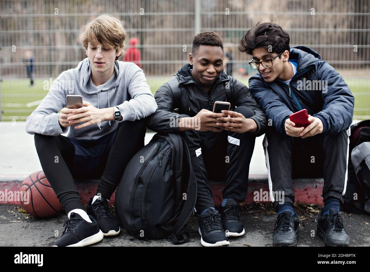 Social media addicted friends using mobile phones while sitting on sidewalk after basketball practice in city Stock Photo