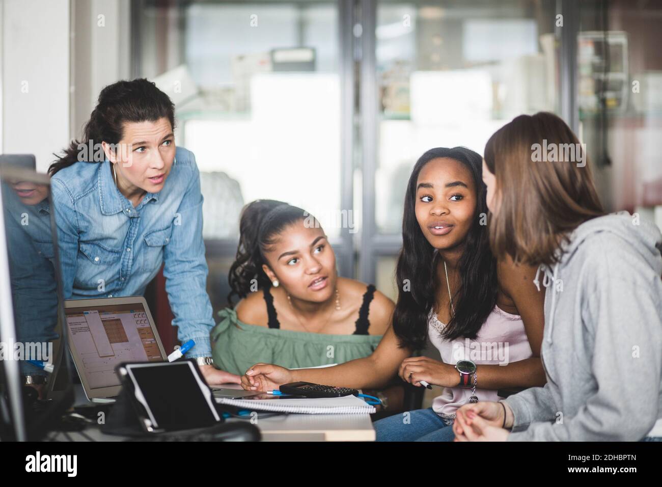 Mature teacher looking at confident female high school students discussing over laptop in computer lab Stock Photo