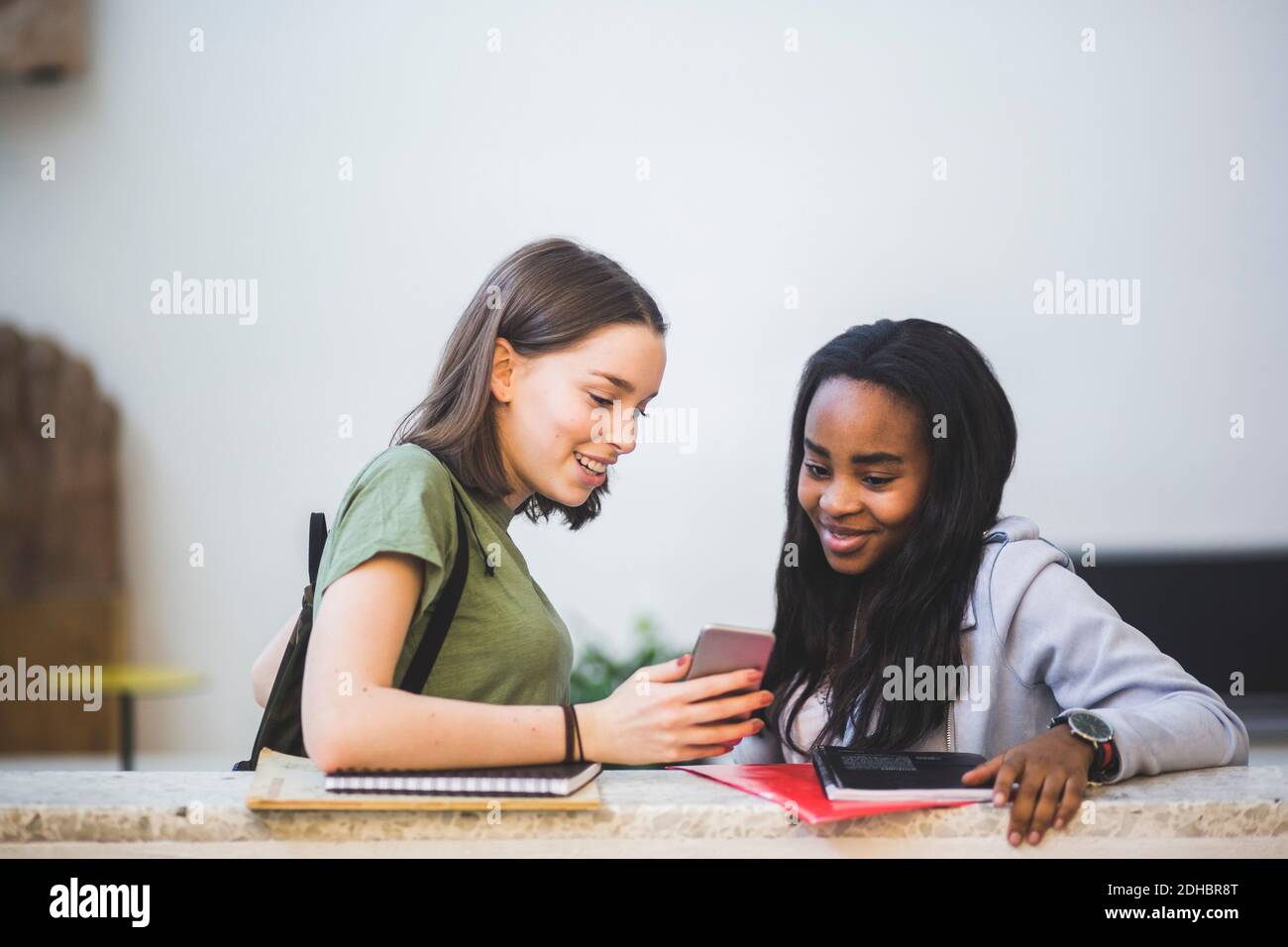 Smiling multi-ethnic female students sharing smart phone while standing in corridor at high school Stock Photo