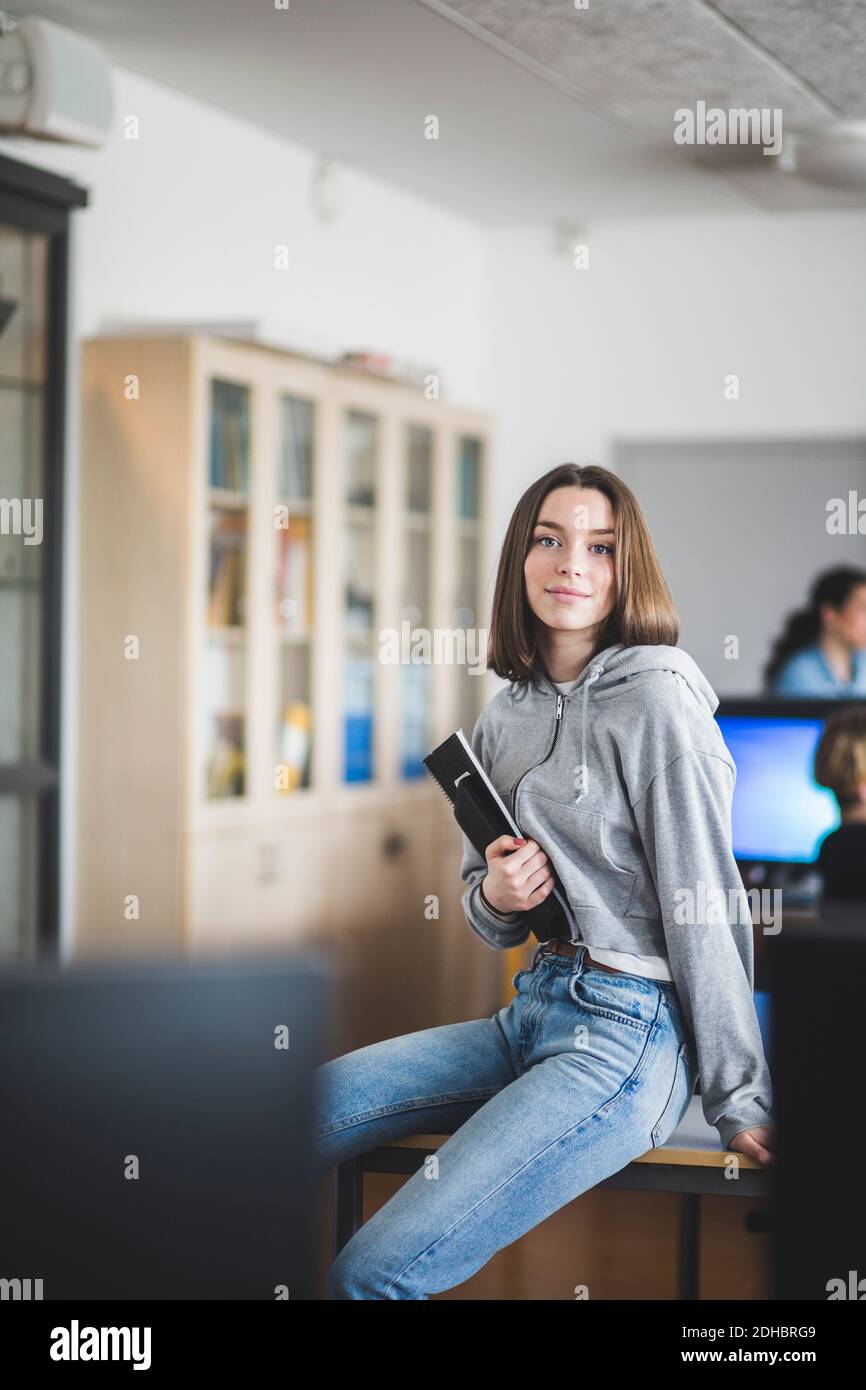 Portrait of confident female student sitting with books on desk in classroom at high school Stock Photo