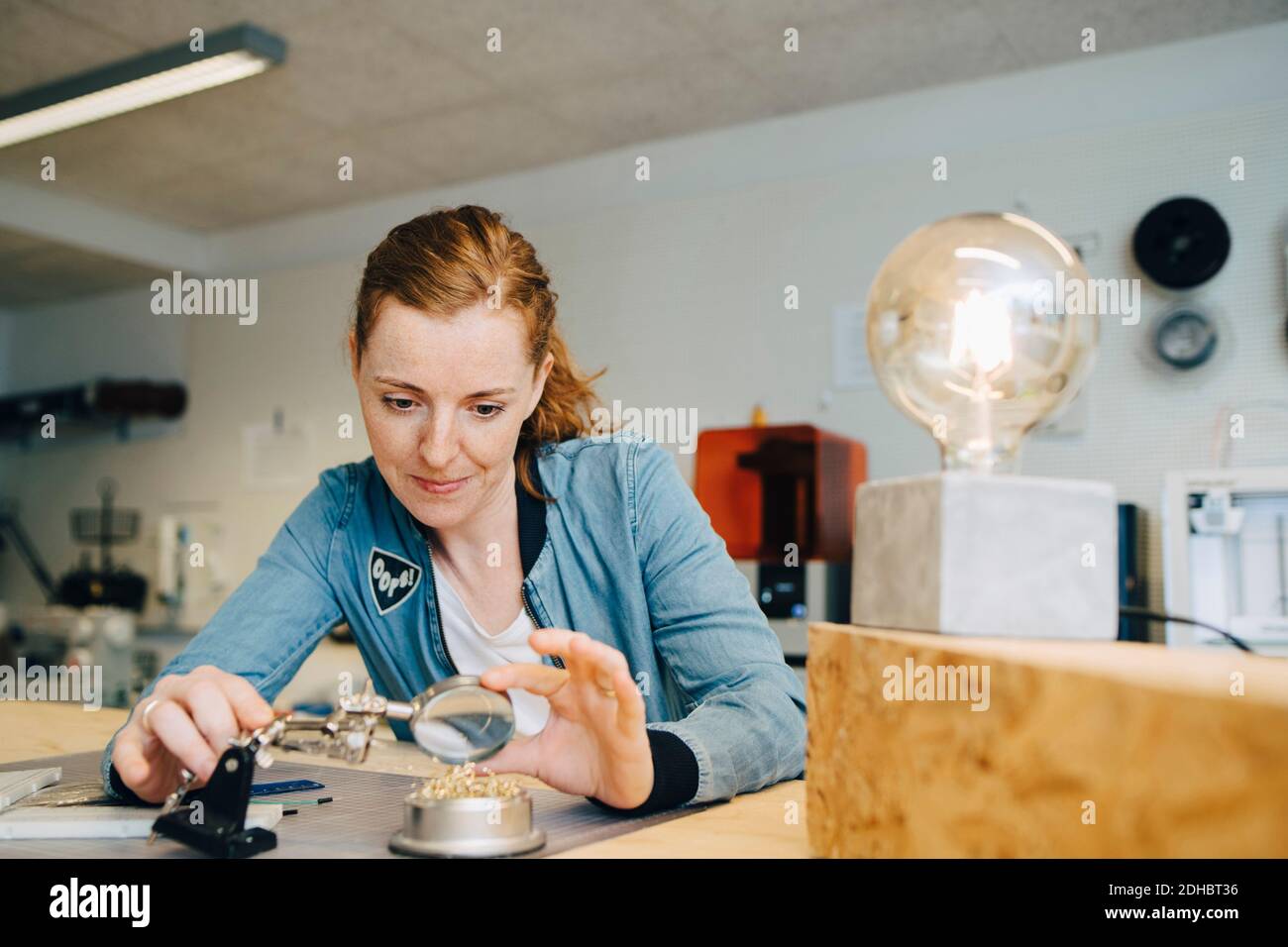 Confident redhead female engineer looking through magnifying glass at workbench in creative office Stock Photo