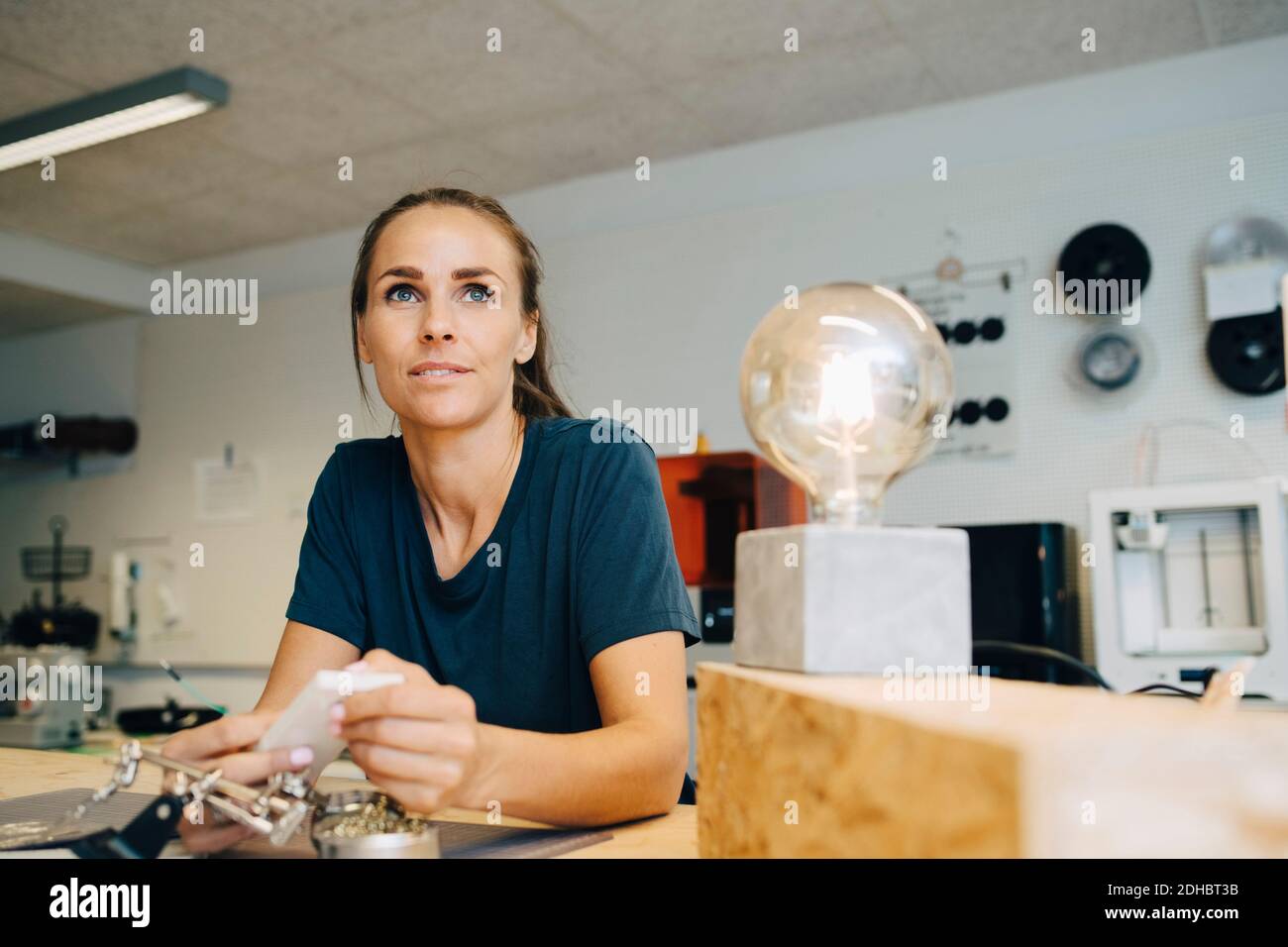 Thoughtful female technician looking away while sitting at workbench in creative office Stock Photo