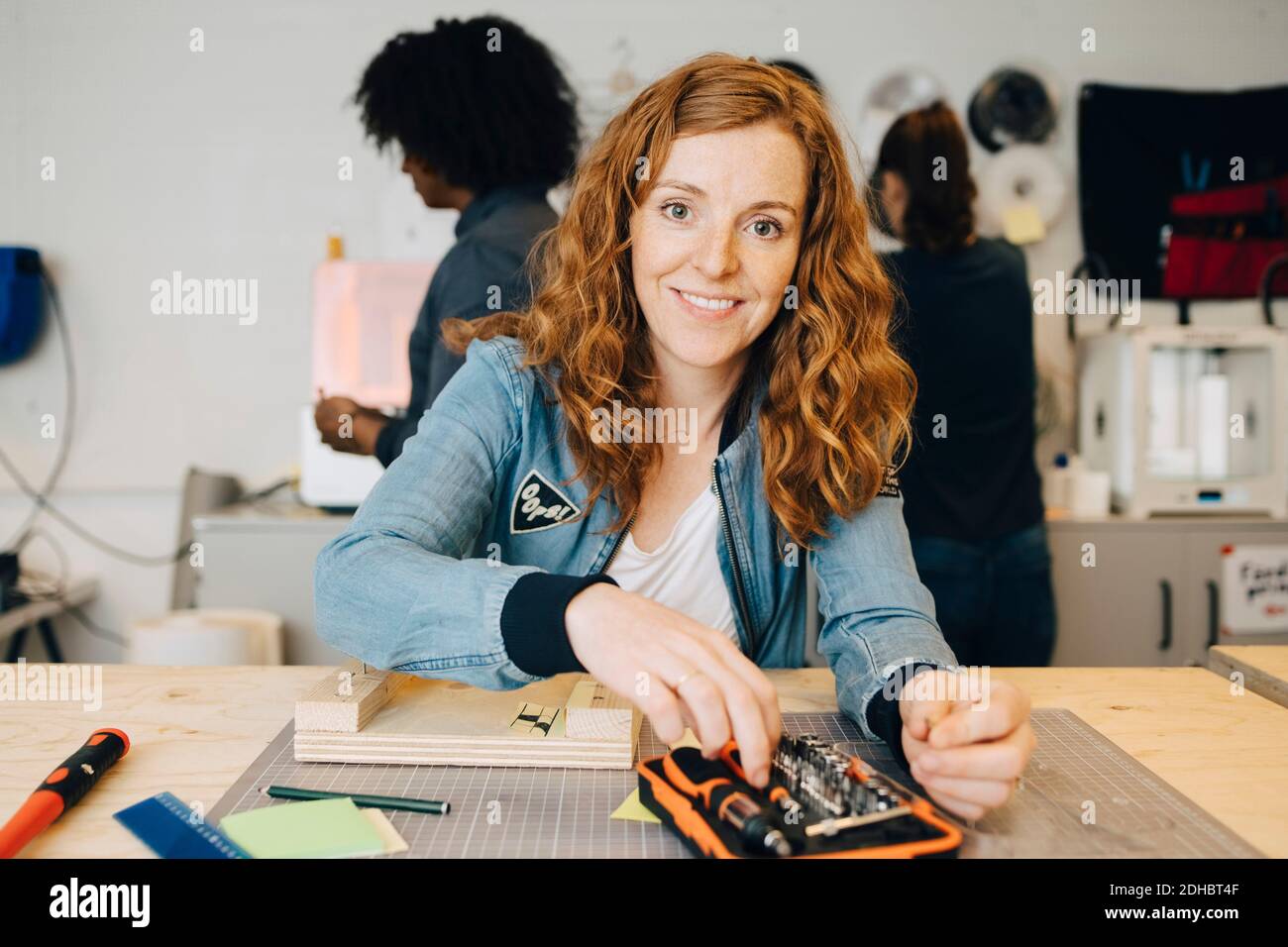 Portrait of smiling redhead female technician sitting at workbench while colleagues working in creative office Stock Photo