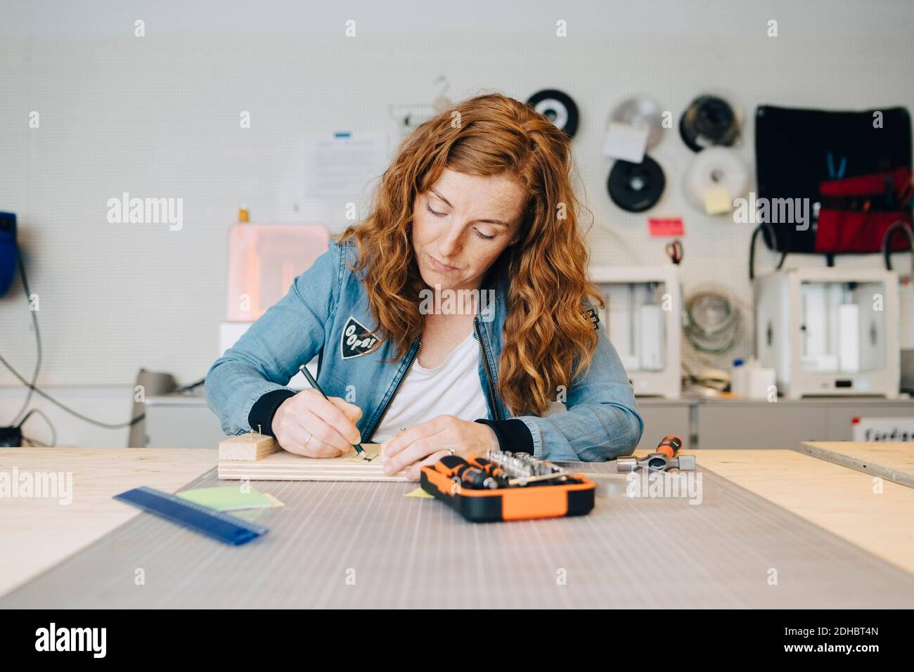 Confident redhead female engineer writing on wood at workbench in creative office Stock Photo