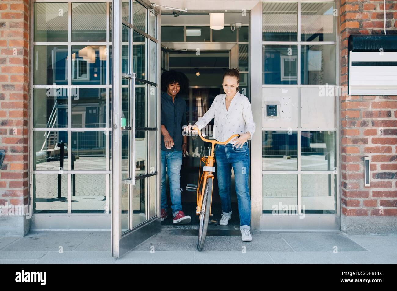Full length of smiling businesswoman holding bicycle while walking with colleague through doorway at office Stock Photo