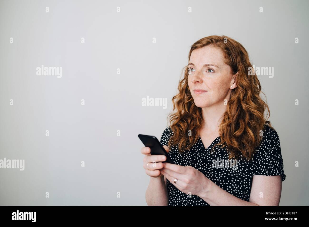 Thoughtful redhead businesswoman standing with smart phone against white wall at creative office Stock Photo
