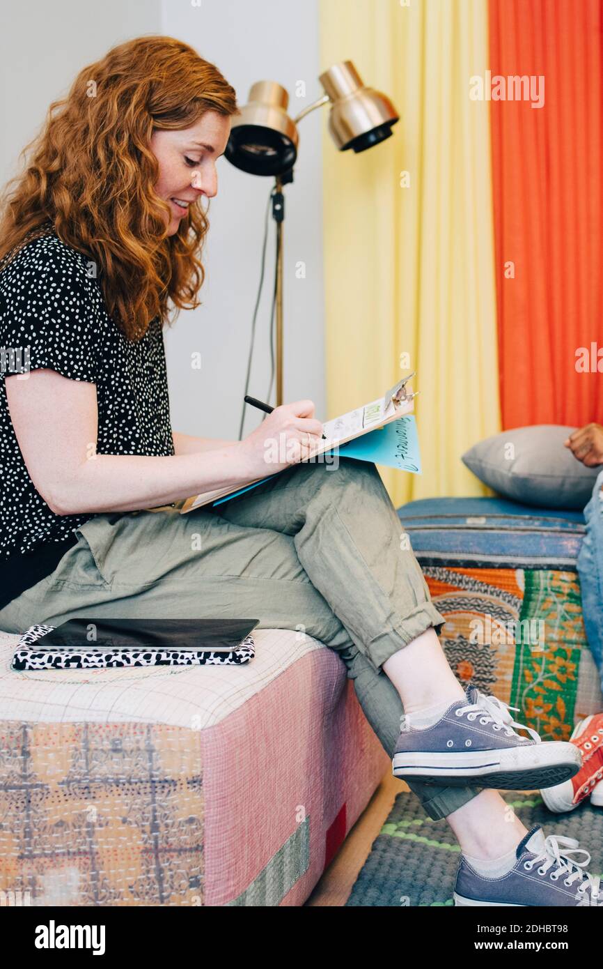 Full length side view of redhead businesswoman writing on clipboard while sitting in lobby at creative office Stock Photo