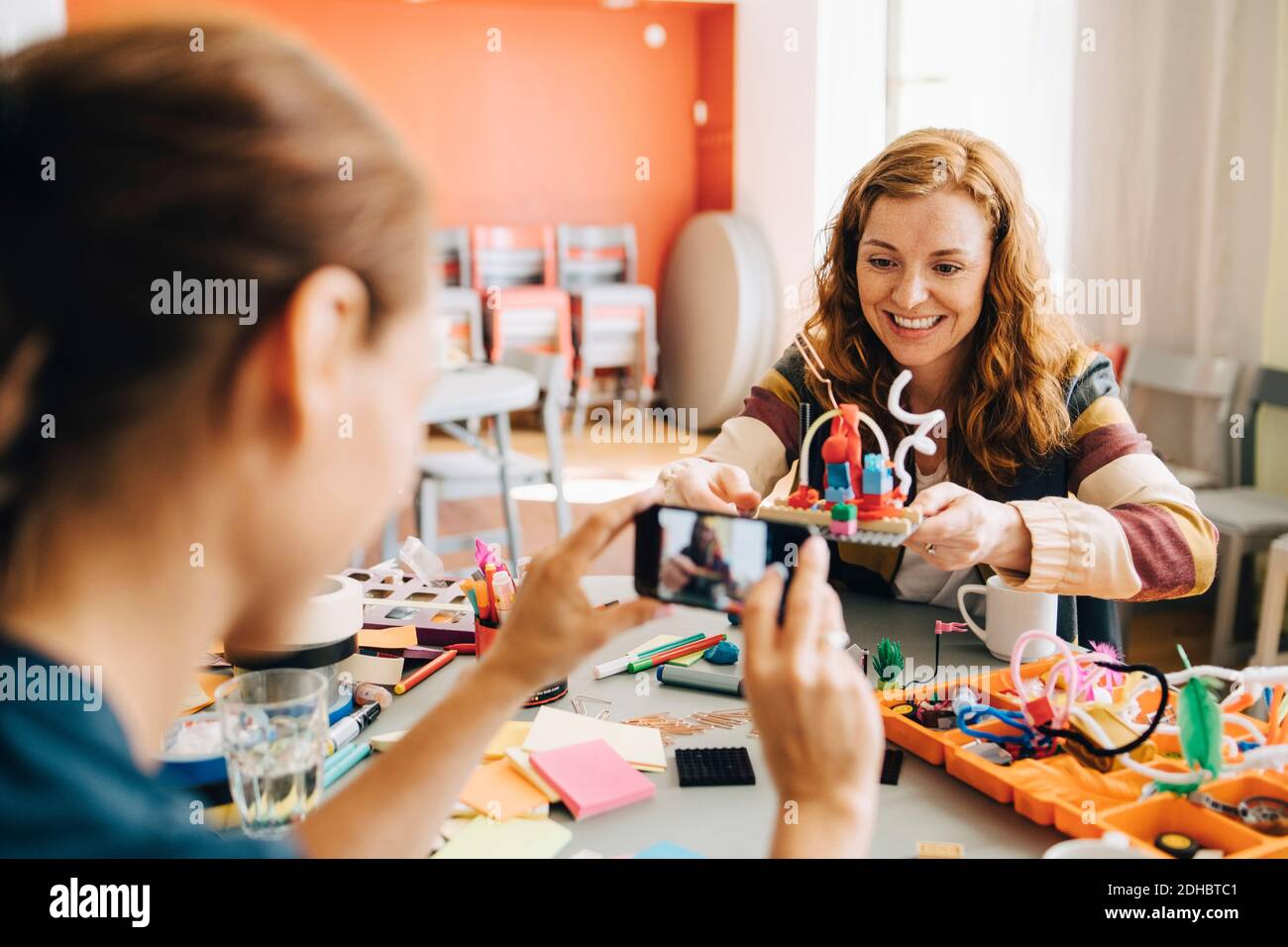 Businesswoman photographing smiling female colleague holding toy at table in creative office Stock Photo