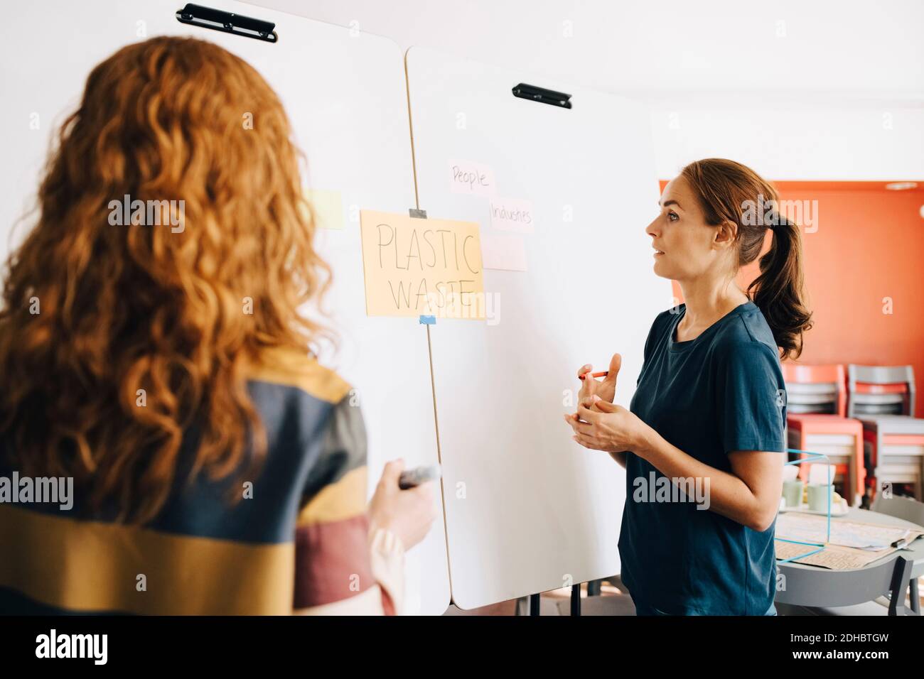 Confident female colleagues discussing plastic management project over whiteboard at creative office Stock Photo