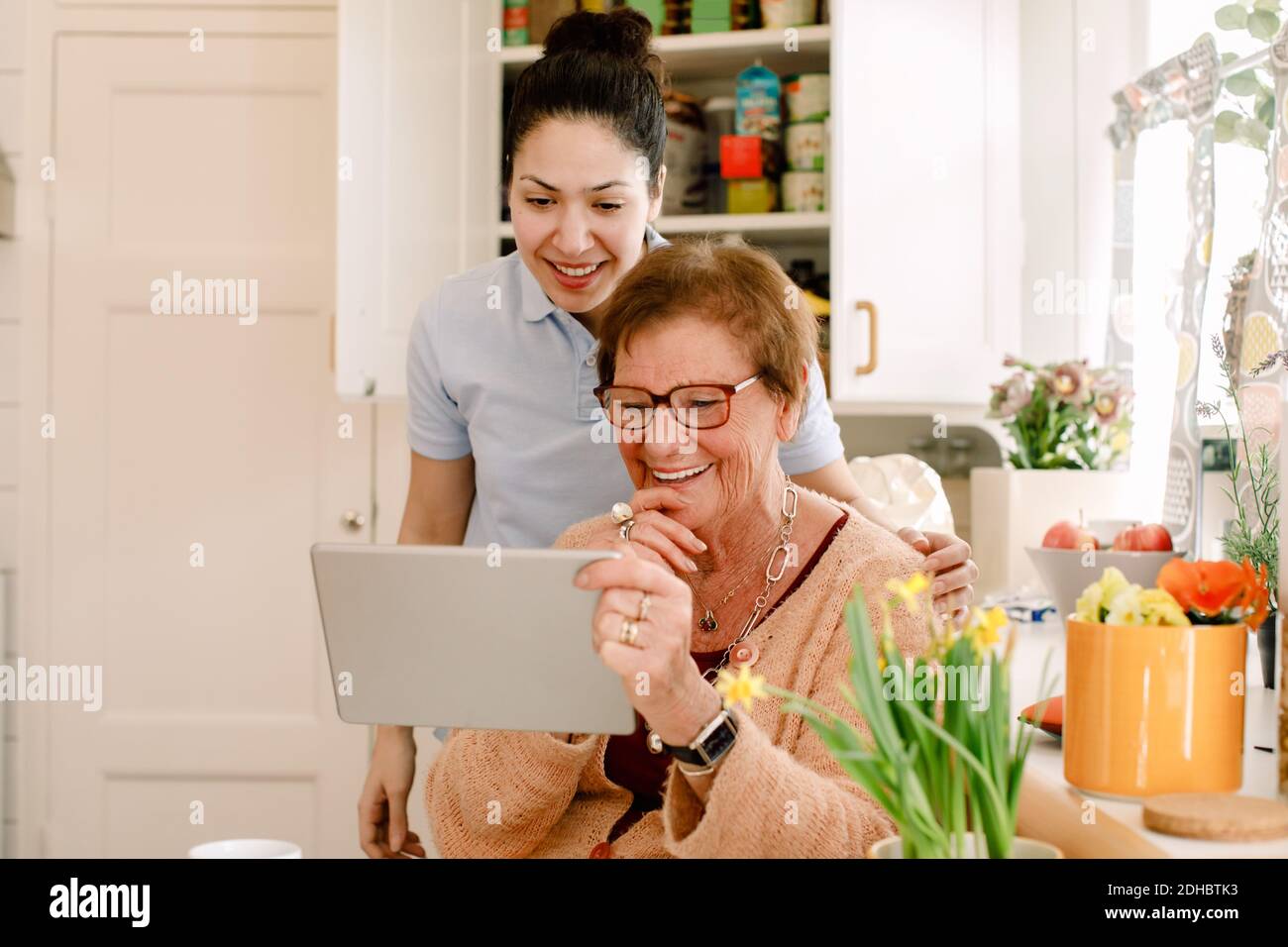 Smiling elderly woman and young female caregiver looking at digital tablet in nursing home kitchen Stock Photo