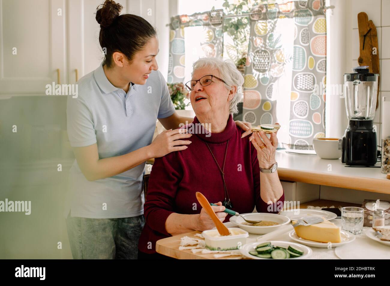 Retired senior woman eating breakfast while talking with female volunteer at nursing home Stock Photo